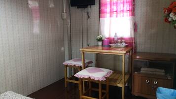 Ein kleines Zimmer mit einem Holztisch und zwei passenden Hocker, einem an der Wand montierten Fernseher, einem Fenster mit rosa Vorhängen und einem Schrank mit blauem Mülleimer.