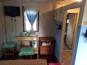 A small room with a wooden table, two green cushioned stools, a cabinet, a television mounted on the wall, and a window with a curtain. - Phaiwan Baansawn Resort (Chai Nat)