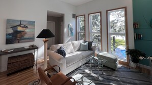 A modern living room with a sofa, a glass coffee table, a ceiling fan, and large windows offering a view of the outdoors. - Stunning Stono River Guesthouse w/ Scenic Water Views &  Pool (Charleston)