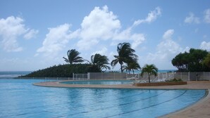Una piscina de borde curvo, rodeada por una valla blanca, palmeras y vistas al océano.