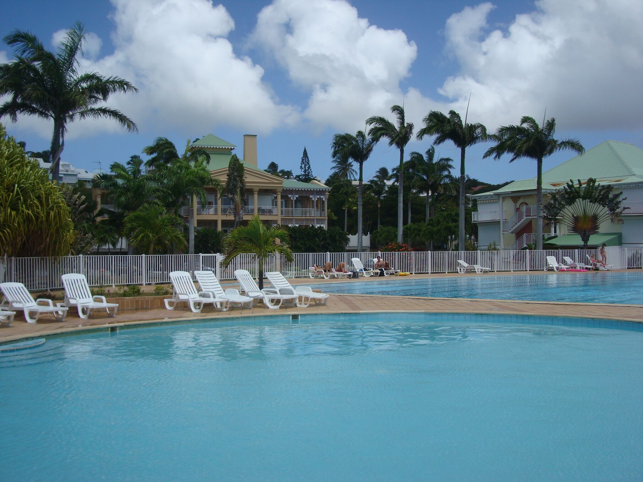 Une piscine entourée de chaises longues blanches, de palmiers et, au loin, d’un bâtiment d’hôtel.