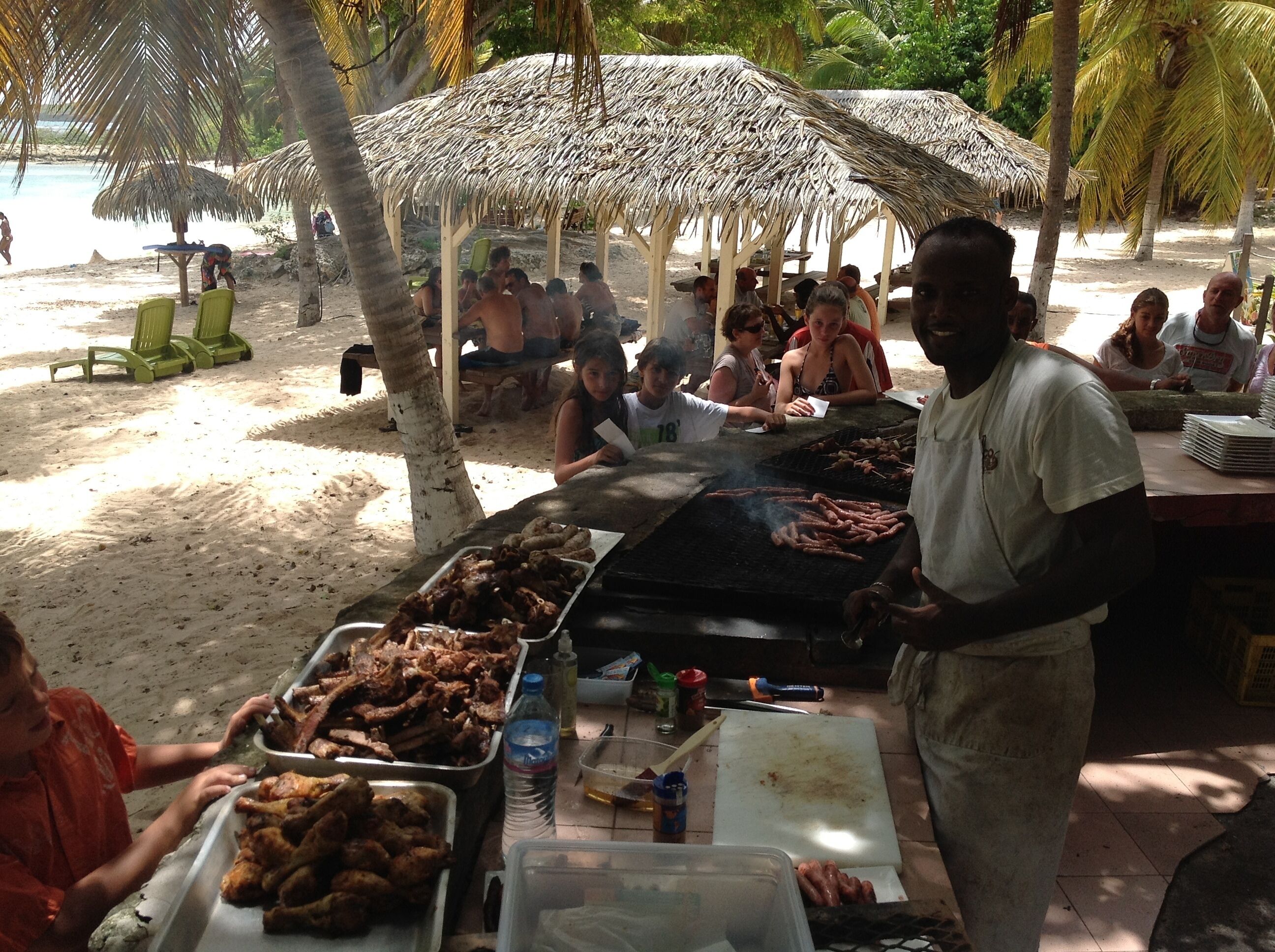 Una barbacoa en la playa con gente reunida alrededor de una parrilla, bajo sombrillas de paja y palmeras al fondo.