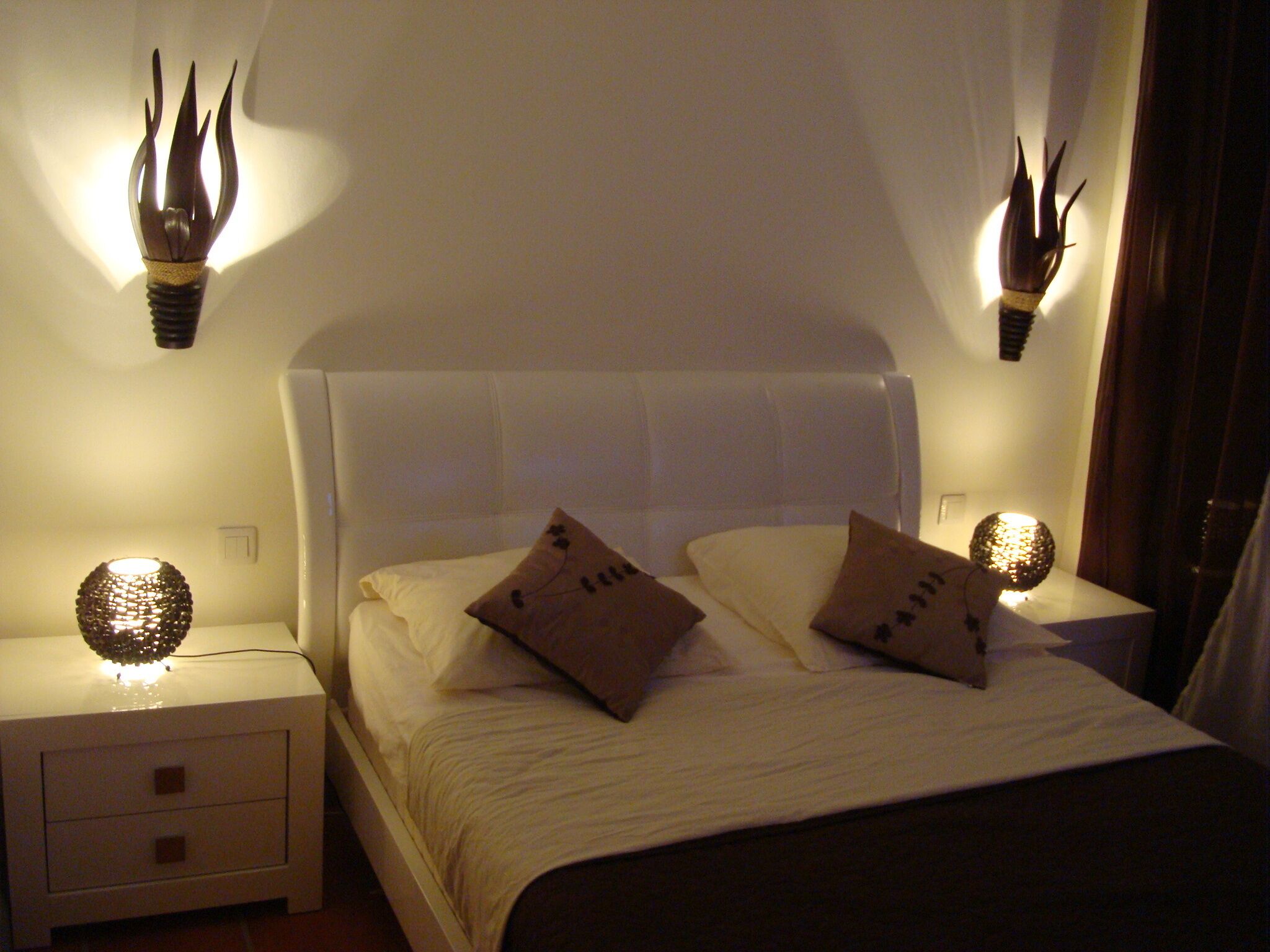 A neatly made bed with two pillows, a white headboard, and two wall-mounted lamps with decorative elements.
