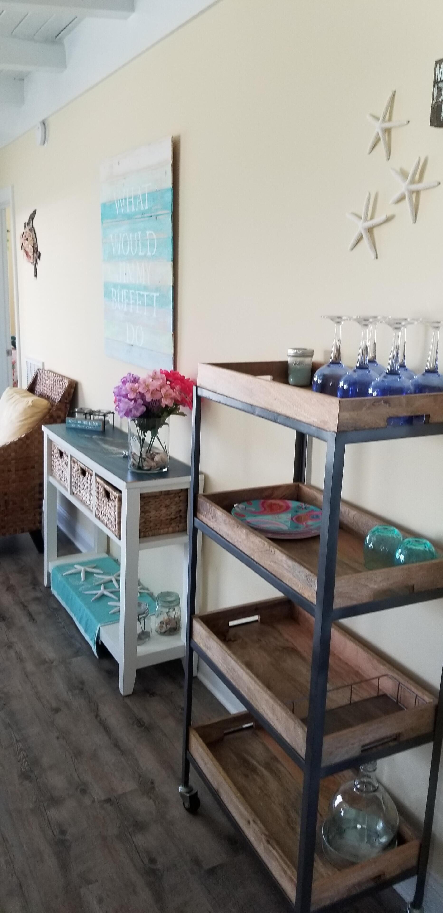 A room with a wooden floor, a white wall with a sign, a wooden table with glassware, and a wicker chair.