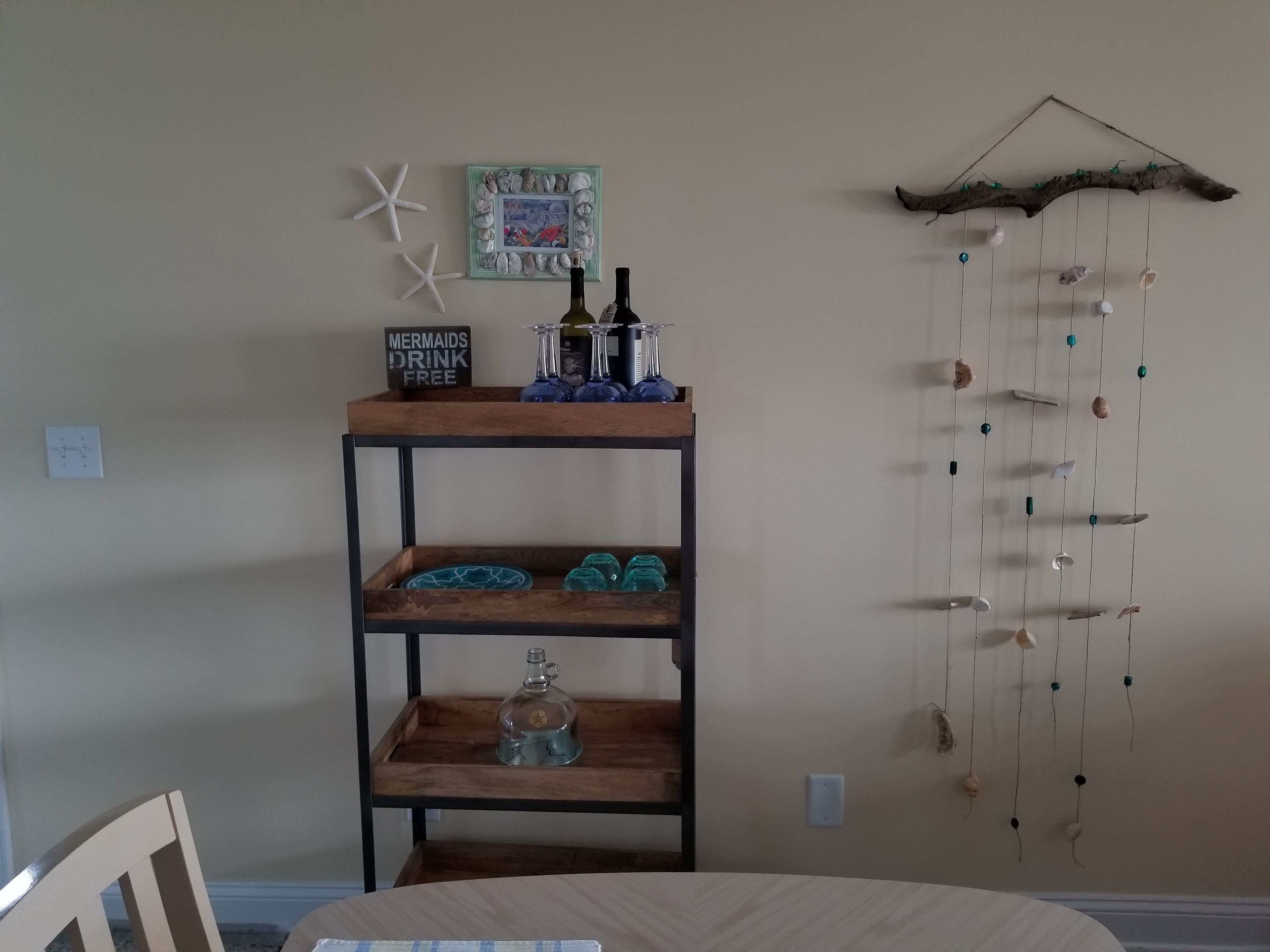Une étagère en bois ornée d’objets décoratifs, une pancarte indiquant « Les sirènes boivent gratuitement » et une décoration murale composée de coquillages et de perles.