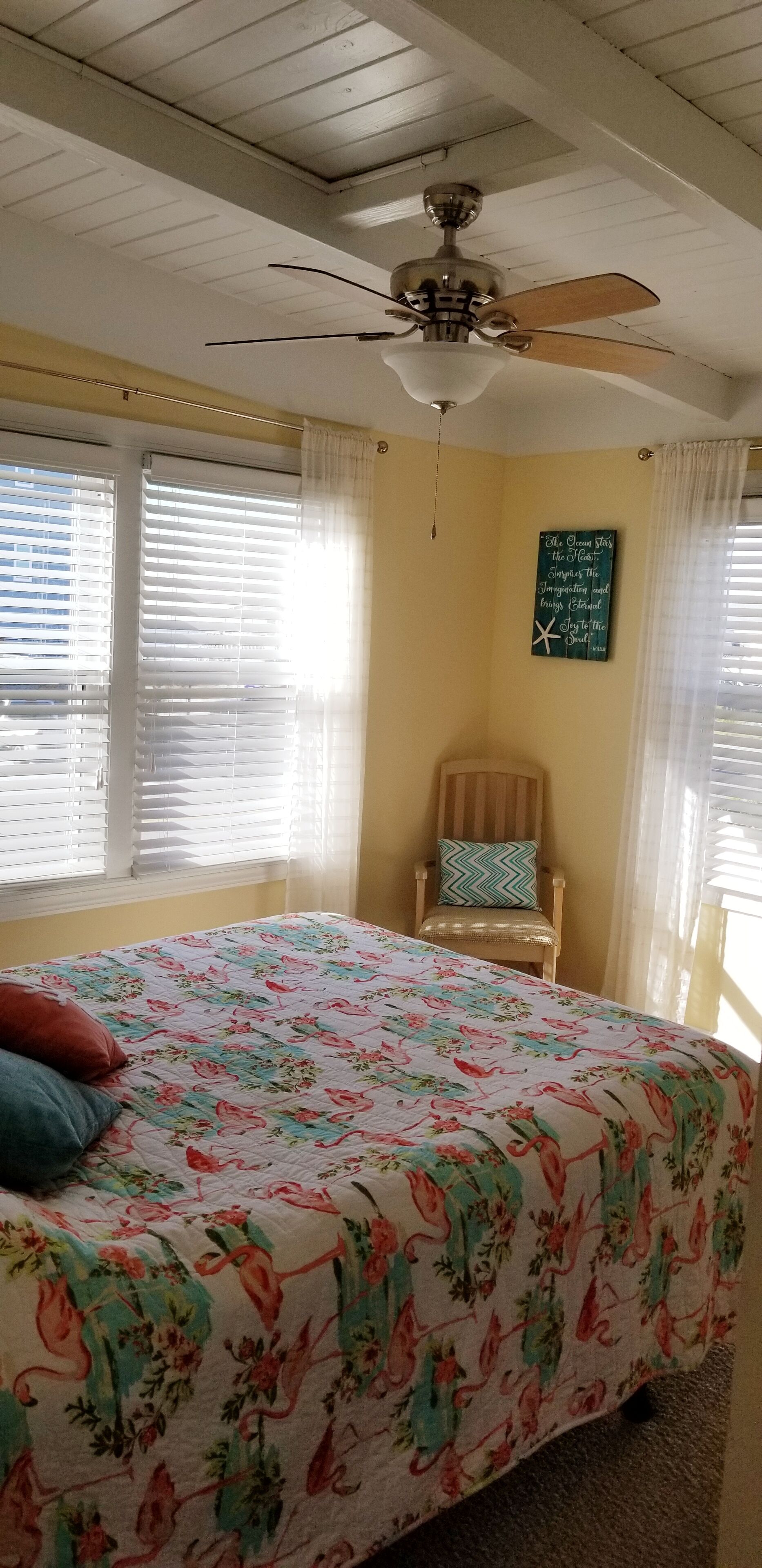 Une chambre à coucher avec un lit, un ventilateur de plafond, une chaise et un mur orné d’une citation inspirante.