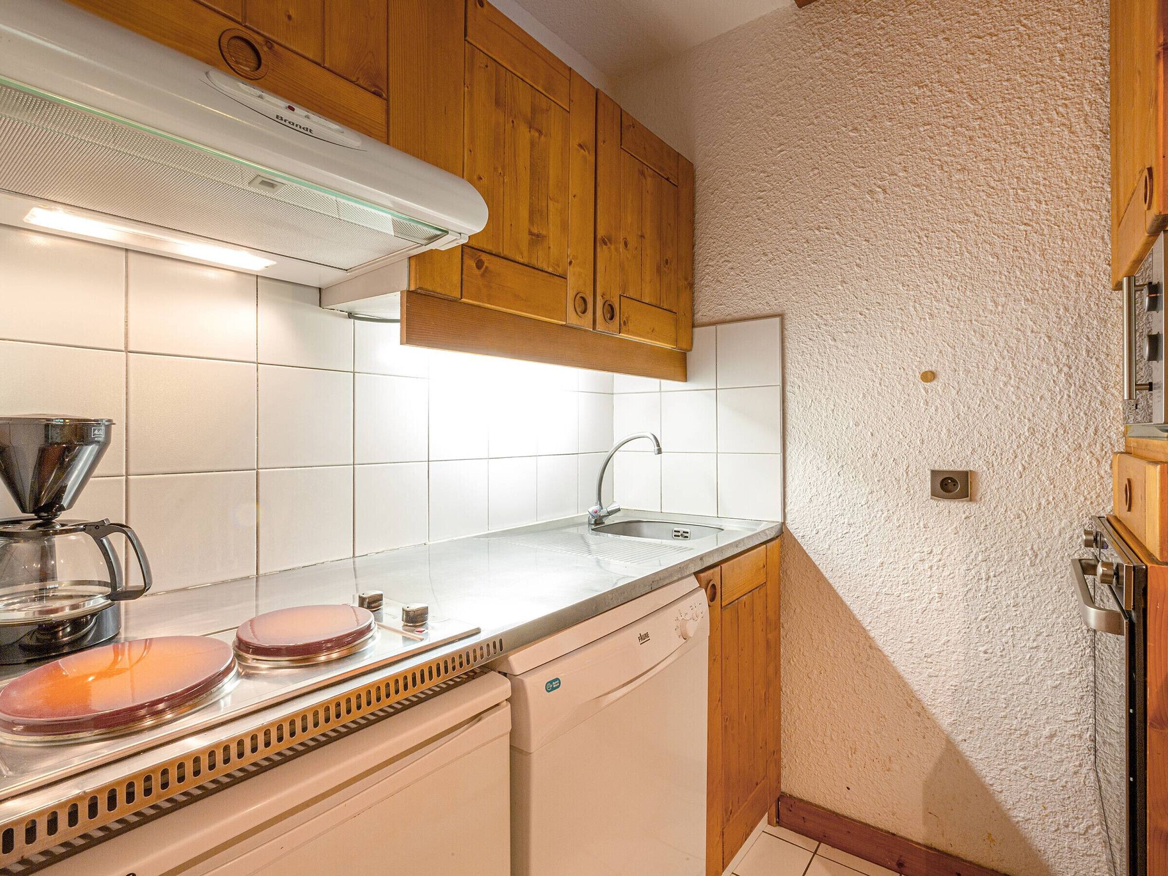 Una cocina compacta con baldosas blancas, armarios de madera, horno empotrado y cafetera.