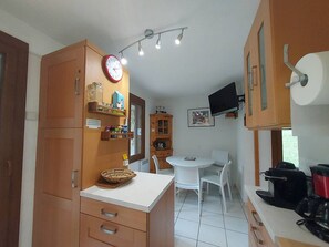 A compact kitchen with wooden cabinets, a white countertop, a dining table with chairs, a clock, and a TV mounted on the wall.