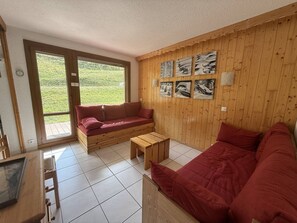A room with a wooden floor, a red sofa, a wooden coffee table, and a view of greenery through a large window.