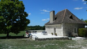 Un bâtiment en pierre avec un toit en tuiles, une cheminée et une terrasse aménagée avec du mobilier d’extérieur.