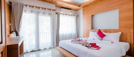 A modern hotel room with a wooden ceiling, a large bed with white bedding and pink accents, a desk with a chair, and a large window with curtains.