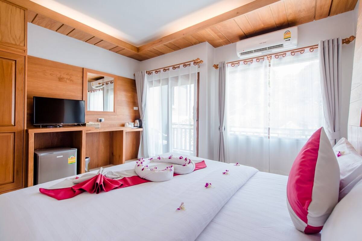 A hotel room with a wooden ceiling, a flat-screen TV, a minibar, a bed with white bedding and pink pillows, and a window with curtains.