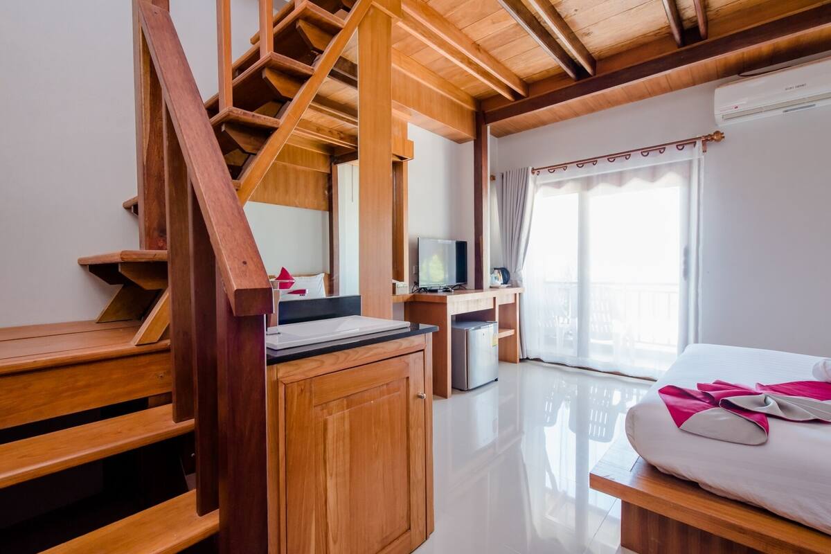A wooden staircase leading to a balcony, a bed with a white and pink blanket, a desk with a computer, and a television.