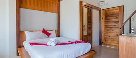 A neatly made bed with white linens and a red accent pillow, a wooden bench at the foot of the bed, and a wooden door with a glass panel.