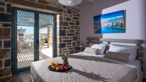 A bedroom with a stone wall, a bed with a quilt, a tray of fruit, and a painting of a coastal scene. - Mandy Luxury Villa by Cretevasion (Kokkini Hani)