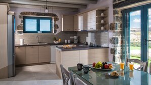 A modern kitchen with a stone accent wall, a large window, and a dining area with a glass table. - Mandy Luxury Villa by Cretevasion (Kokkini Hani)