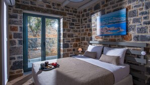 A bedroom with stone walls, a bed with a beige bedspread, a nightstand with a lamp, and a painting of a coastal scene. - Mandy Luxury Villa by Cretevasion (Kokkini Hani)