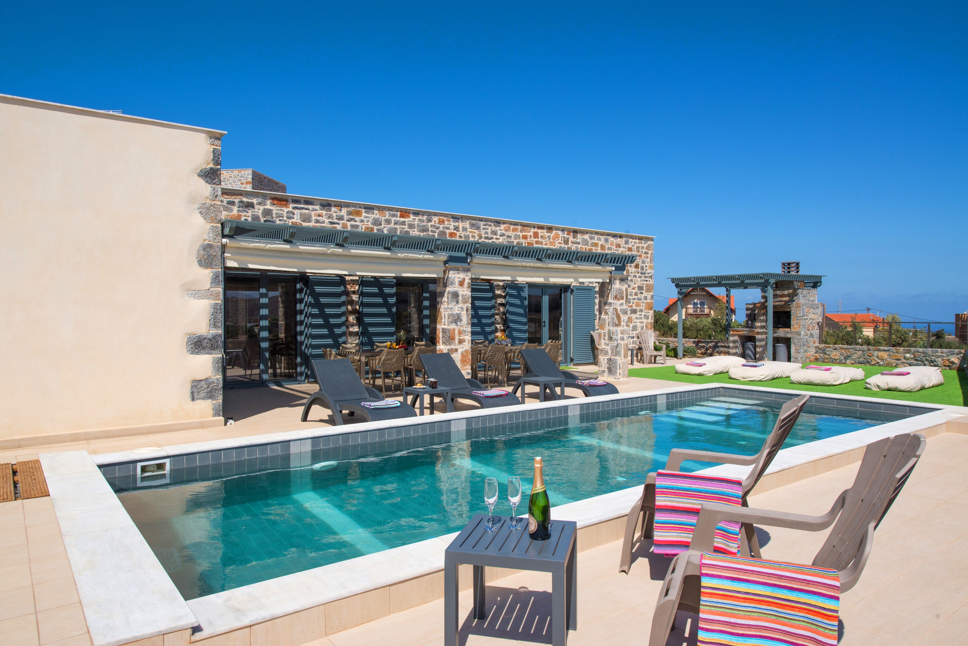 A poolside area with lounge chairs, a table with a bottle and glasses, and a stone building with a covered patio.