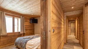 Un dormitorio con paneles de madera, una cama, un banco, un televisor montado en la pared y un pasillo que conduce a otra habitación.
