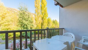 Un balcón con una mesa y sillas blancas, con vistas a un jardín con árboles y una montaña al fondo.