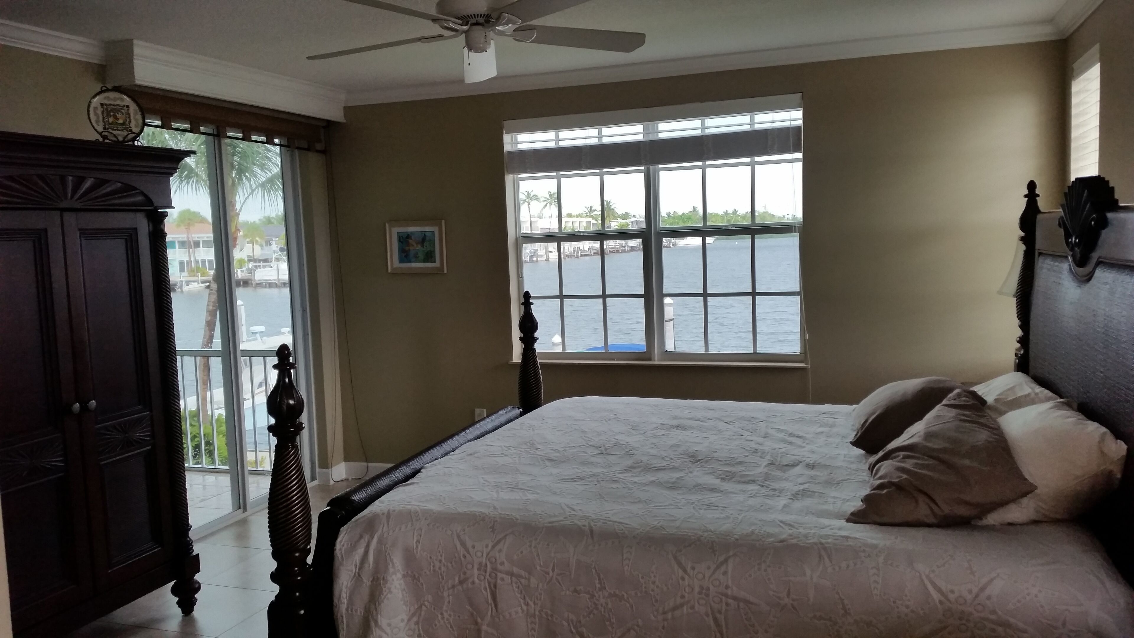 A bedroom with a bed, a window with a view of water, a ceiling fan, and a wooden cabinet.