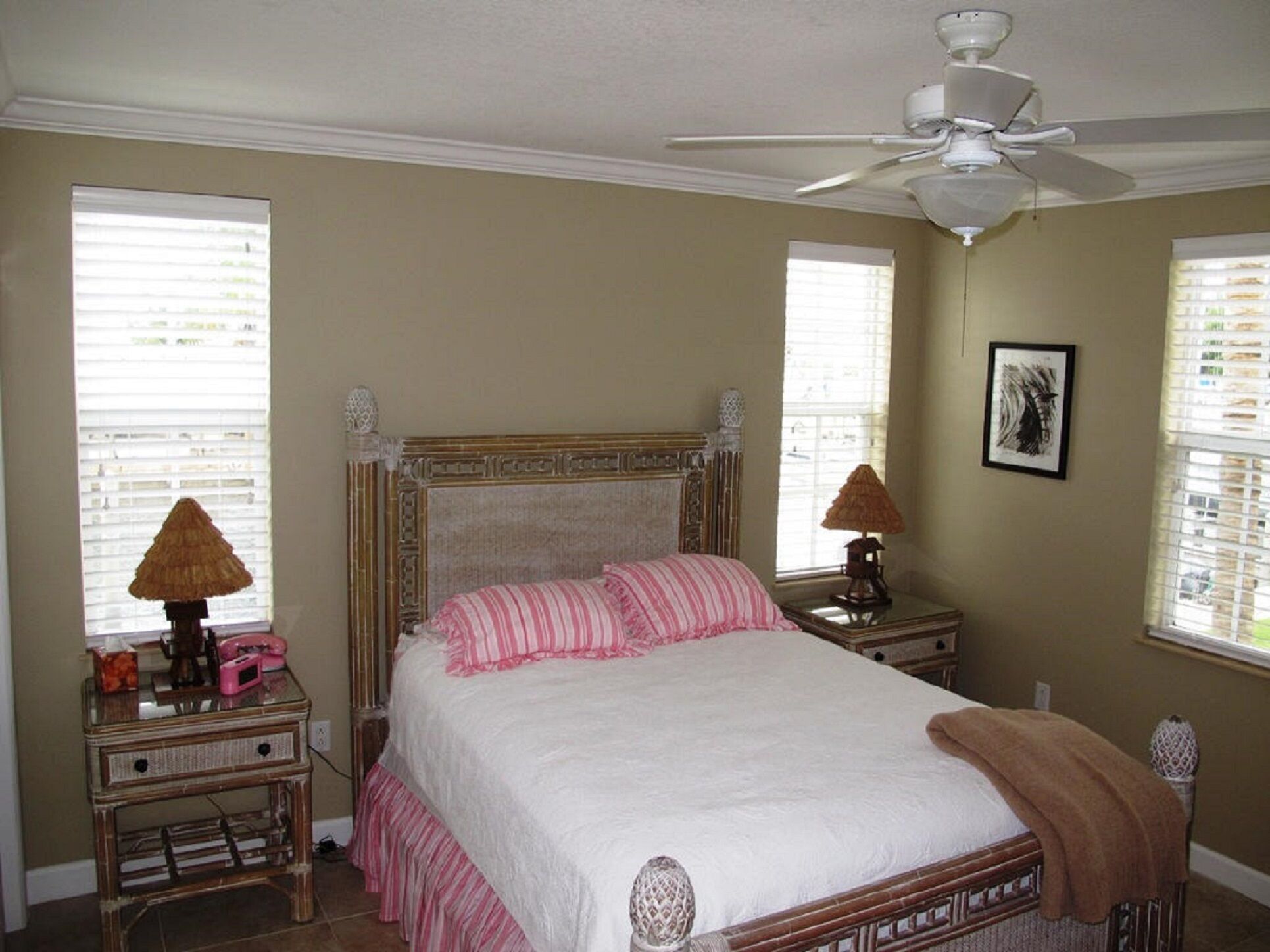 A bedroom with a bed, two bedside tables, a ceiling fan, and windows with blinds.