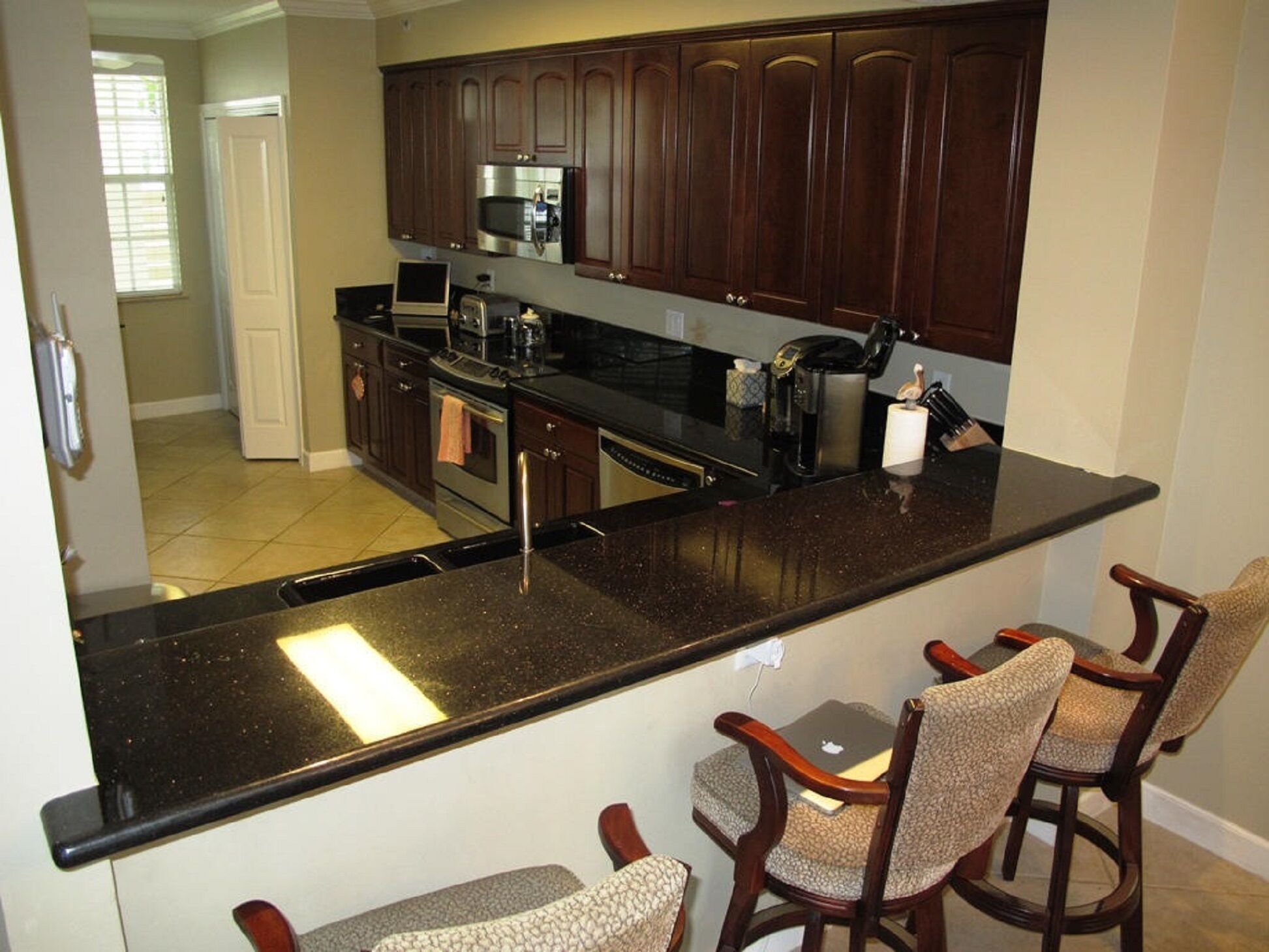 A kitchen with dark wood cabinets, a black granite countertop, and a coffee maker. There are two bar stools with cushioned seats.