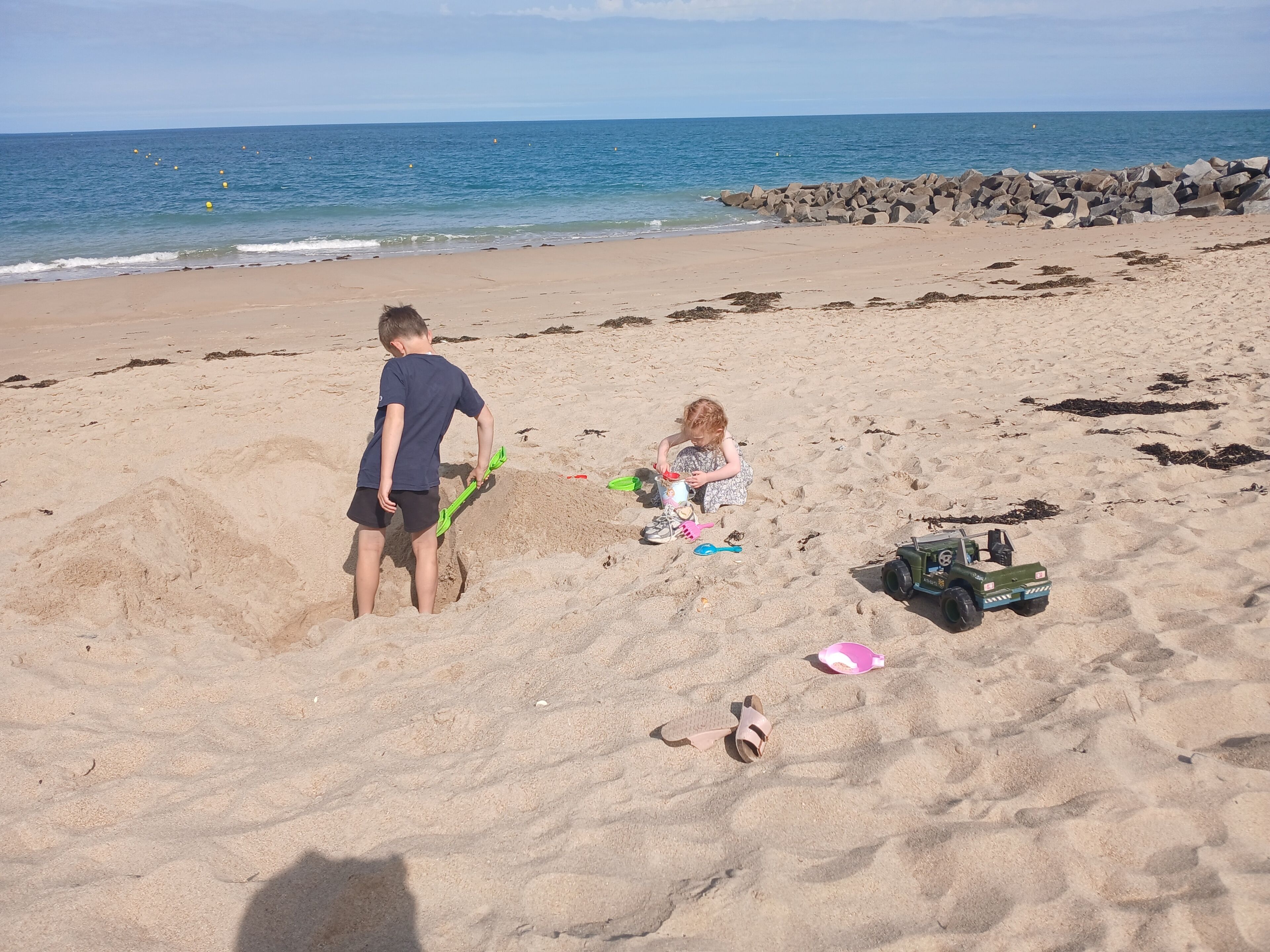 Une plage de sable avec une digue rocheuse, un enfant et un adulte qui jouent, et une voiture jouet.