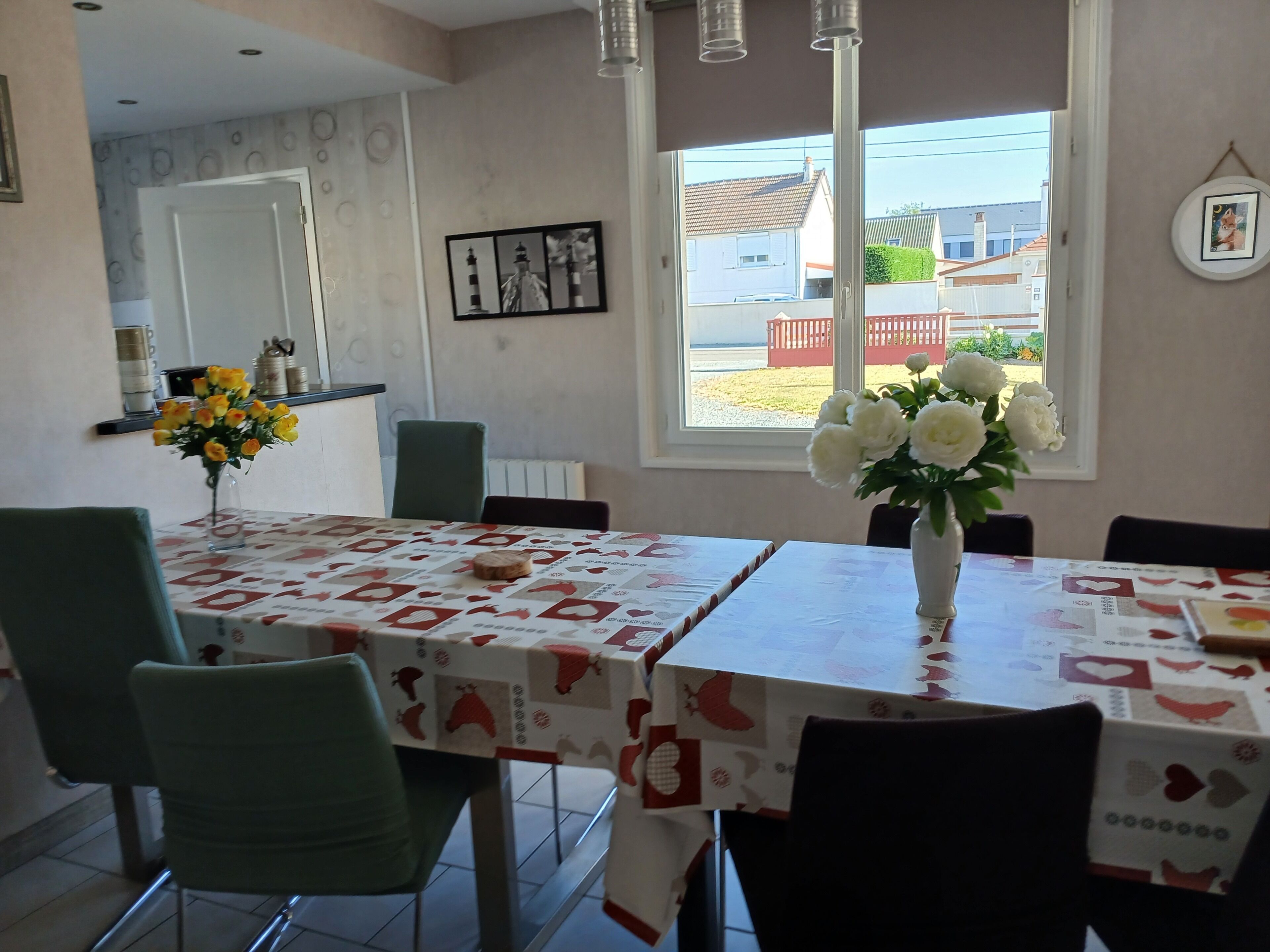 A dining area with a table set for two, chairs, a vase with flowers, and a window with a view of houses.
