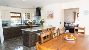 A modern kitchen with a central island, wooden dining table, and chairs. There is a bowl of fruit on the table.