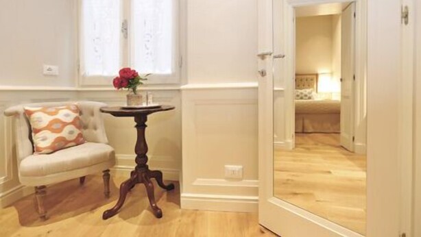 A room with a wooden floor, a white armchair with a patterned cushion, a small round table with a vase of flowers, and a mirror reflecting a bedroom with a bed and a nightstand. - SantaCroce Junior Luxury Suite (Firenze)
