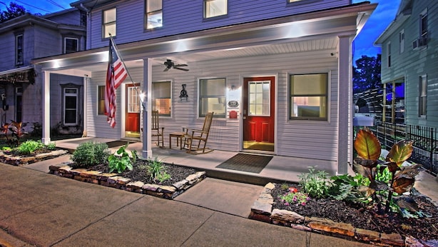 A two-story house with a covered porch, a red door, and a small garden area with plants and a birdbath. - ❤️ Perfect for traveling nurses! - Wifi, AC, 4-6 minute commute to Hershey Med (Hummelstown)