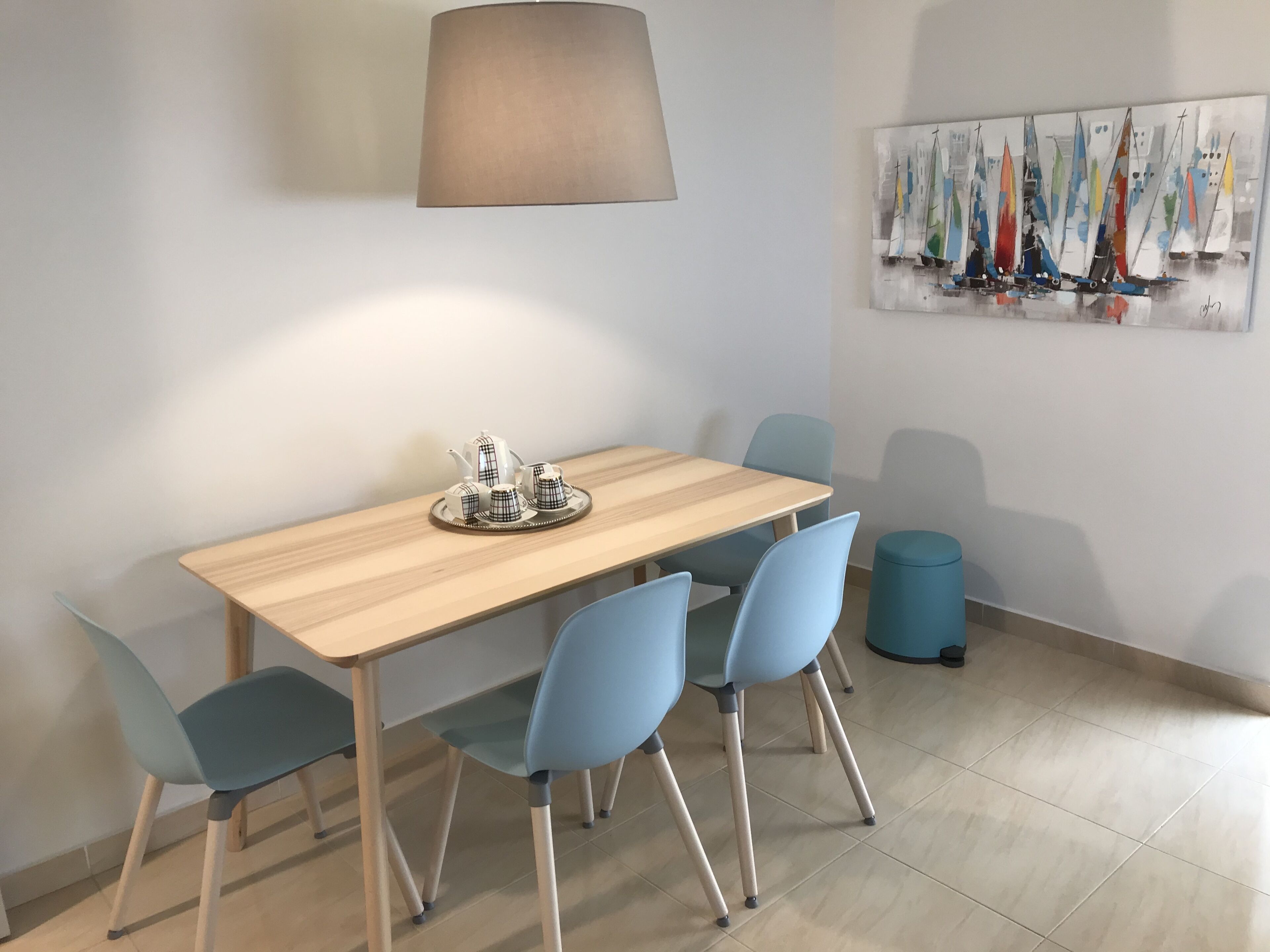 Een eethoek met een houten tafel, vier stoelen, een lamp en een schilderij van zeilboten aan de muur.