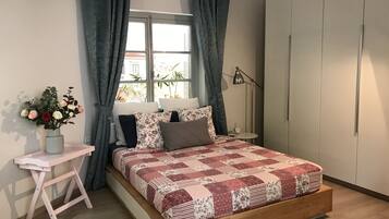 A bedroom with a wooden bed, a small desk, a vase of flowers, and a window with curtains.