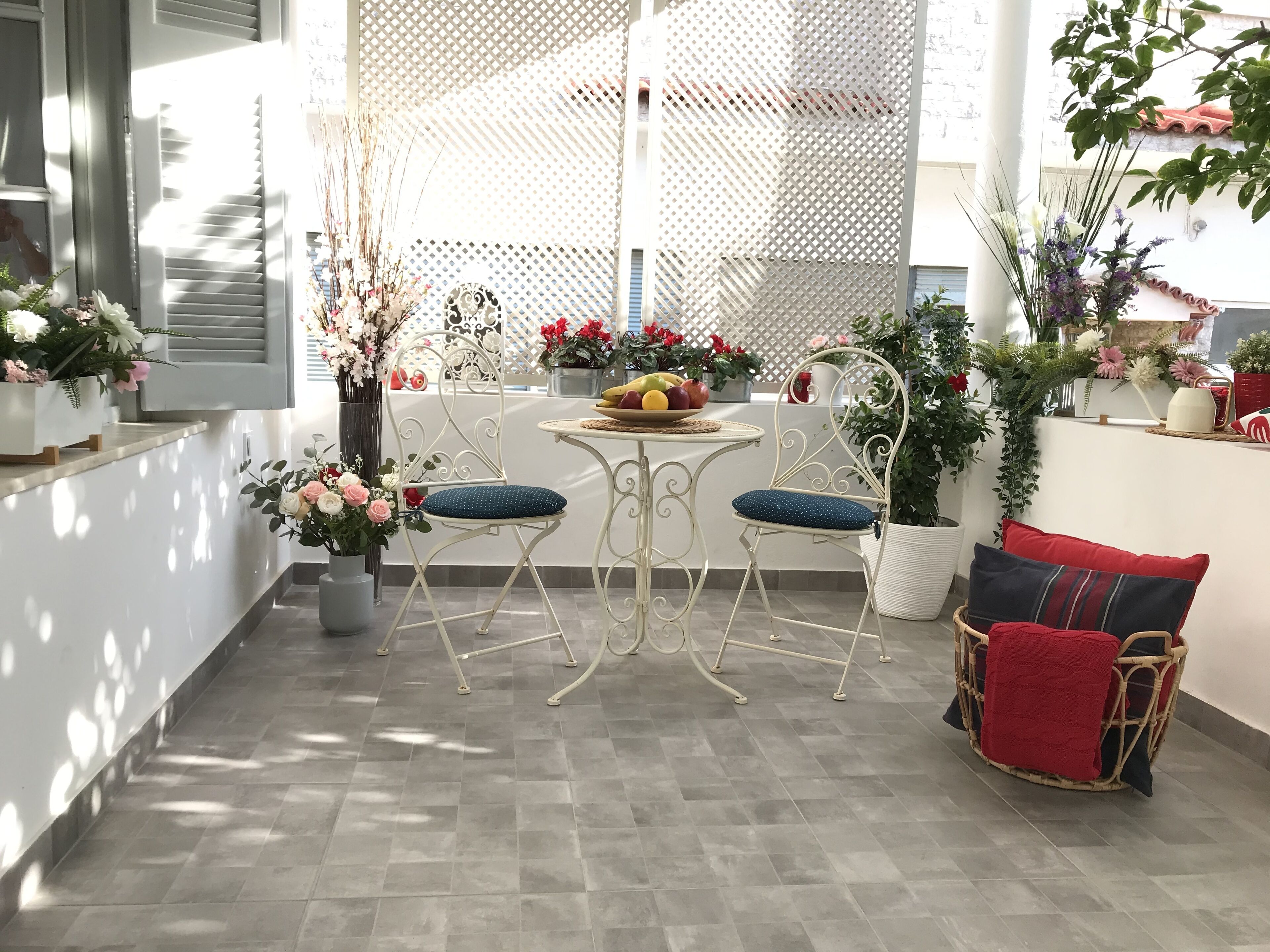 A sunlit patio with a white metal table and two chairs, surrounded by potted plants and a wicker sofa with red cushions.