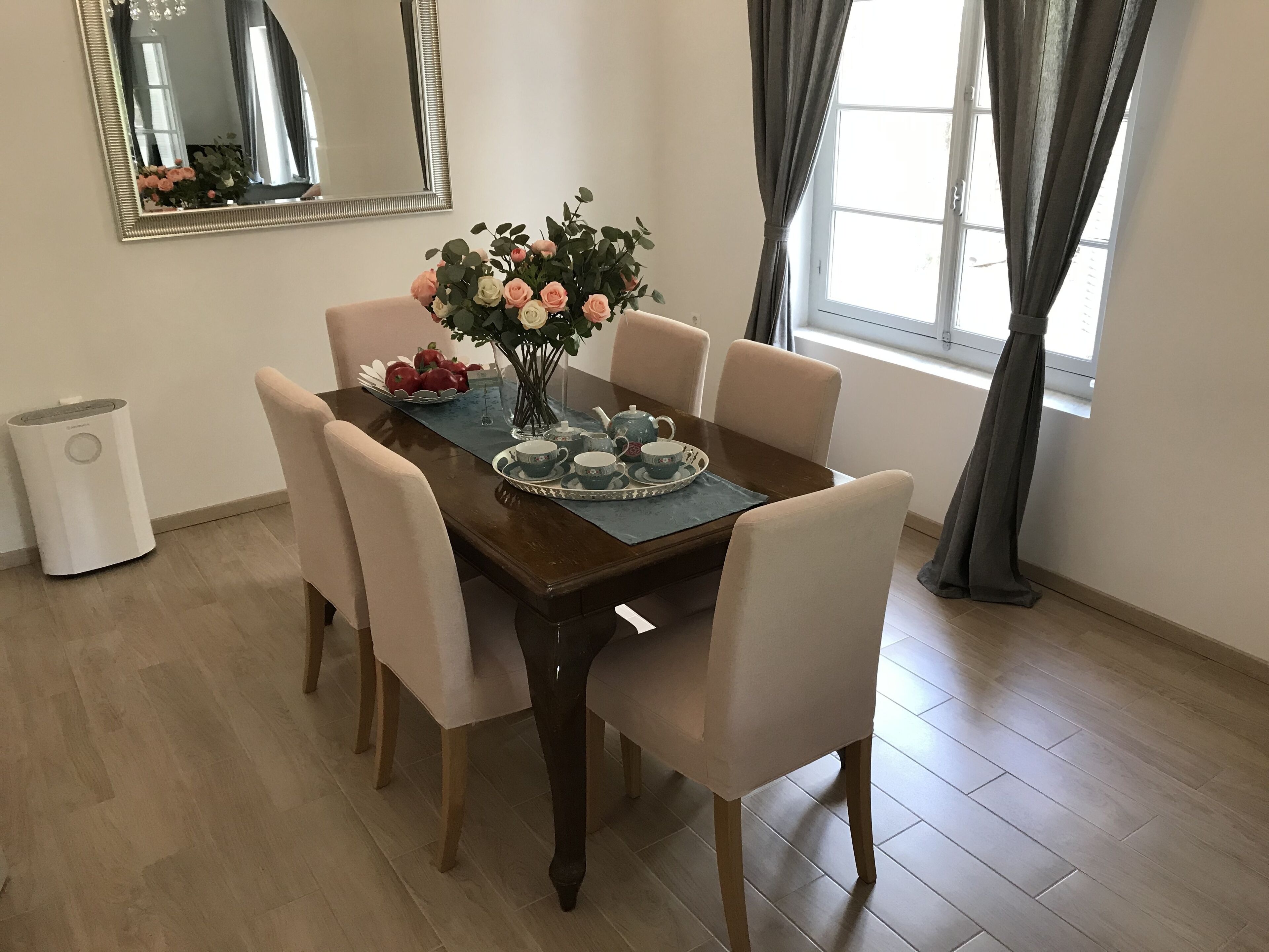A dining area with a wooden table, six chairs, a vase of flowers, and a tray with teacups and apples.