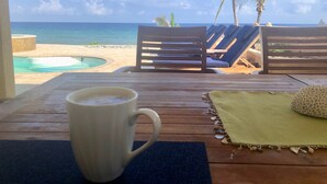 Una mesa de madera con una taza blanca, un mantel verde y una concha decorativa. Al fondo, sillones de madera y una piscina con vistas al océano y palmeras.