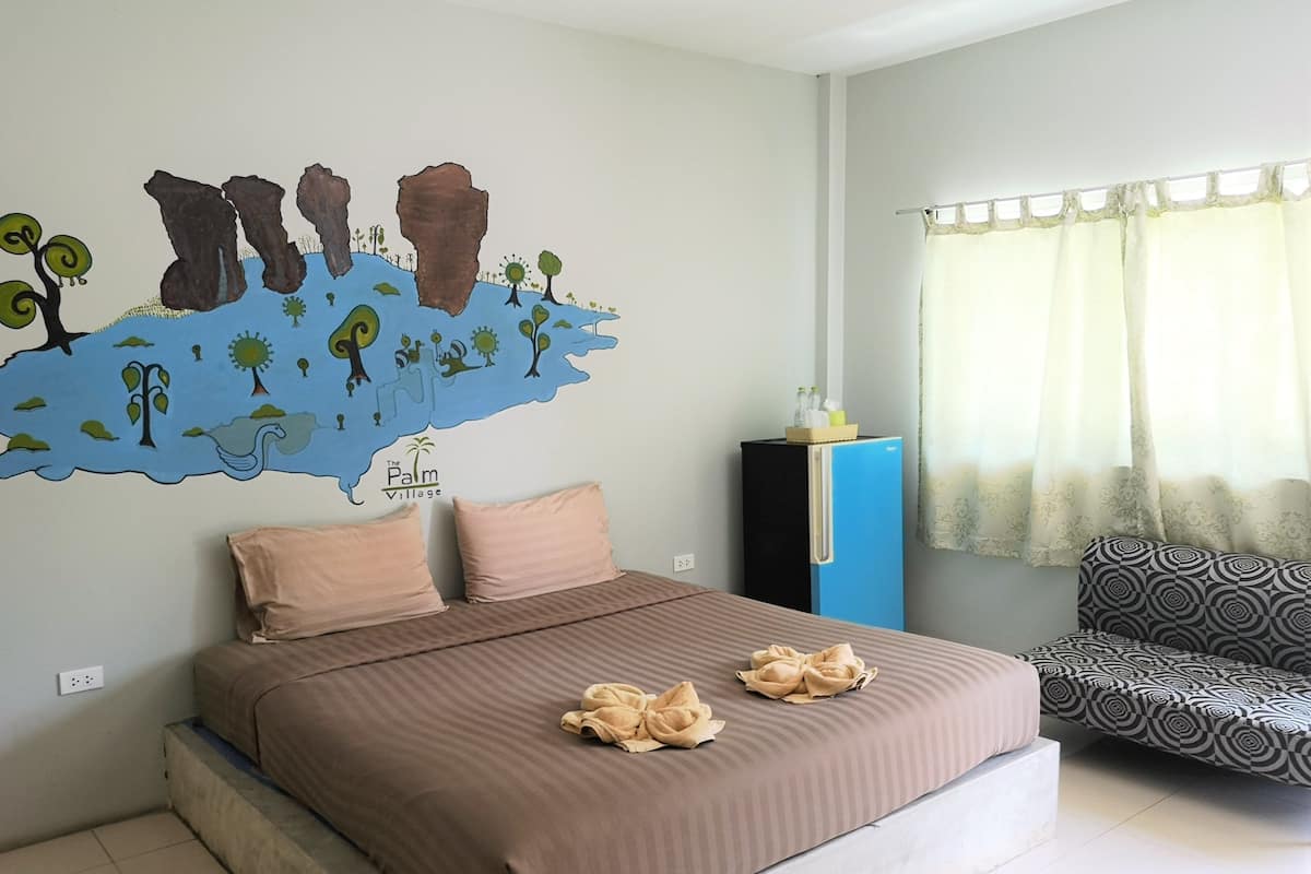 A bedroom with a bed, two pillows, a wall mural, a small refrigerator, and a patterned chair.
