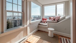A bright room with large windows, a built-in bench with cushions, a white toilet, and a wooden floor.