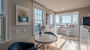 A modern living room with a round wooden table, two blue armchairs, a sofa with patterned cushions, and a view of the city through large windows.
