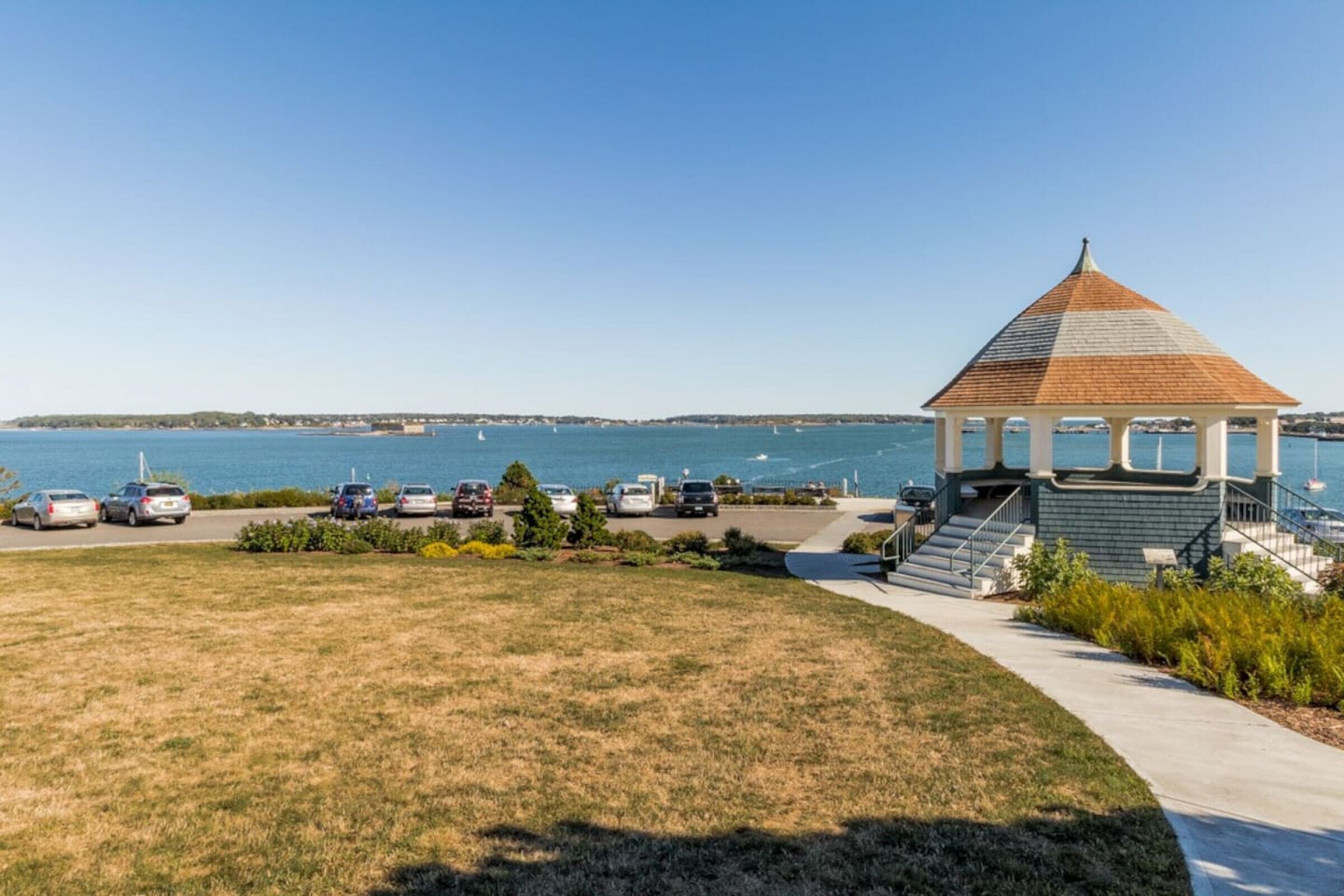 A gazebo with a tiled roof overlooking a parking area and a body of water.