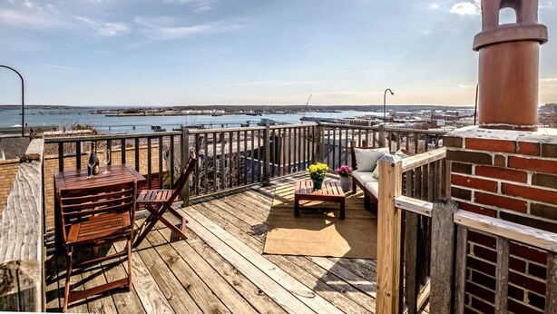 A wooden deck with a brick wall and a chimney, overlooking a marina with boats and a distant cityscape.