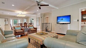 A spacious living room with leather sofas, a patterned ottoman, a dining area with chairs, a ceiling fan, and a TV on a cabinet. - Spacious Isle of Palms Beach House with Private Pool, One Block To The Beach (Isle of Palms)