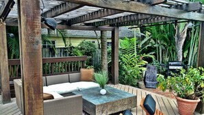 A wooden deck with outdoor furniture, including a sofa and chairs, surrounded by lush greenery and a barbecue grill. - VOTED BEST FORT LAUDERDALE LOCATION! WALK TO SHOPS, RESTURAUNTS, & BEACH. (Ft Lauderdale)