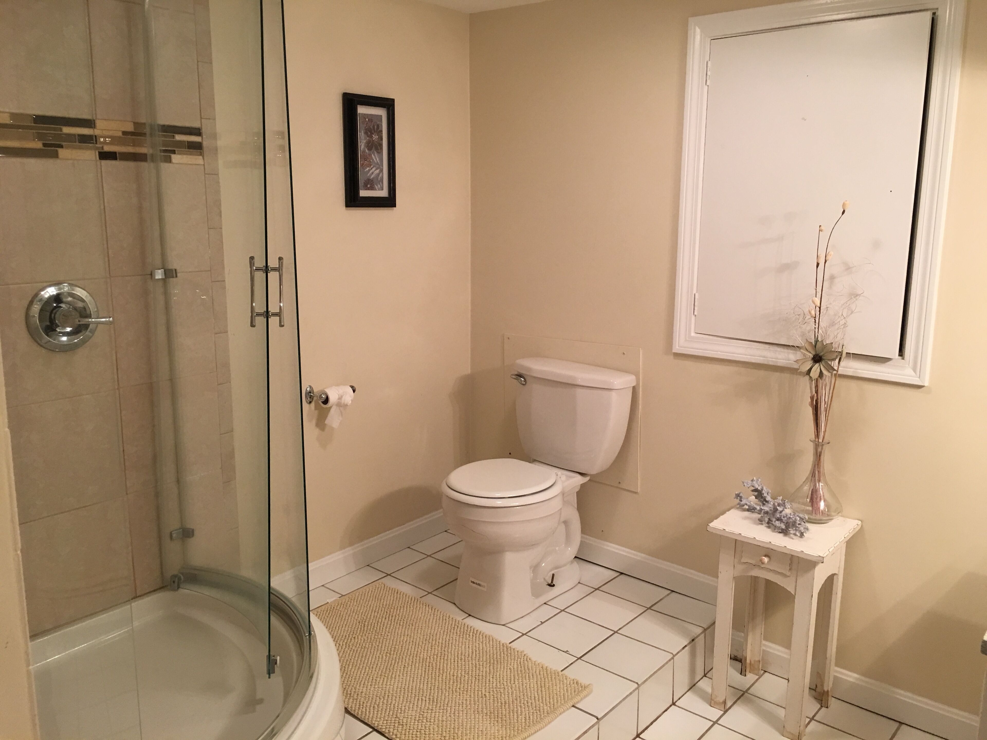 A bathroom with a glass shower enclosure, a toilet, a small white vanity with a vase, and a framed picture on the wall.