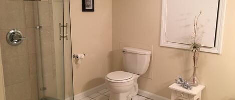 A bathroom with a glass shower enclosure, a toilet, a small white vanity with a vase, and a framed picture on the wall.
