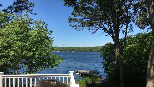 Une terrasse dotée de meubles en osier, donnant sur un lac, entourée d’arbres et bordée d’un quai.