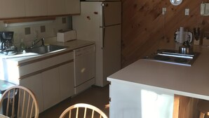 A kitchen with wooden beams, white cabinets, a refrigerator, a stove, and a sink. There are chairs and a table in the dining area. - Rustic and cozy Walk In-Walk Out Ski Condo (Carrabassett Valley)