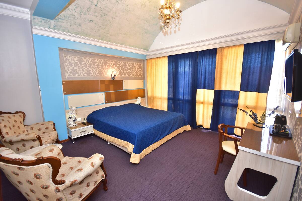 A hotel room with a large bed, two armchairs, a desk with a chair, a vanity with a mirror, and a chandelier.
