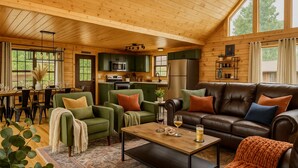 A cozy cabin interior with a dining area, kitchen, and living room furnished with leather sofas and green chairs.